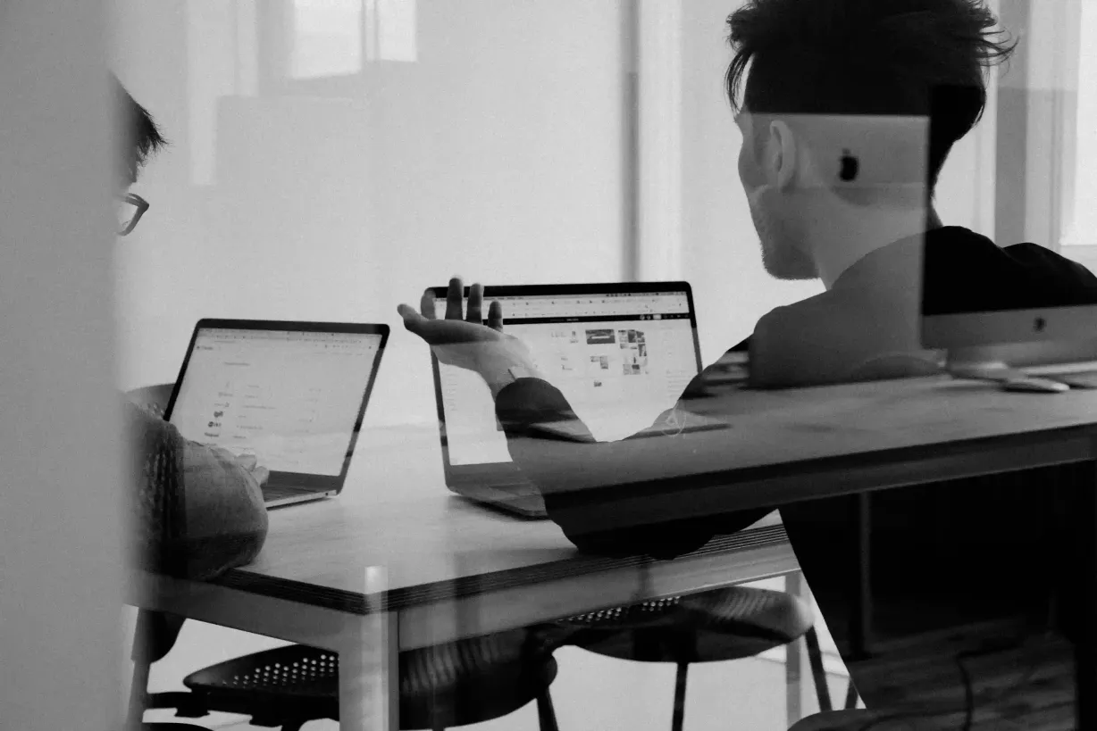 Zwei Menschen sitzen mit Ihren Laptops an einem Tisch und tauschen sich aus.